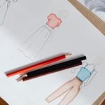 Fashion sketches on paper with red and black colored pencils on a wooden surface.